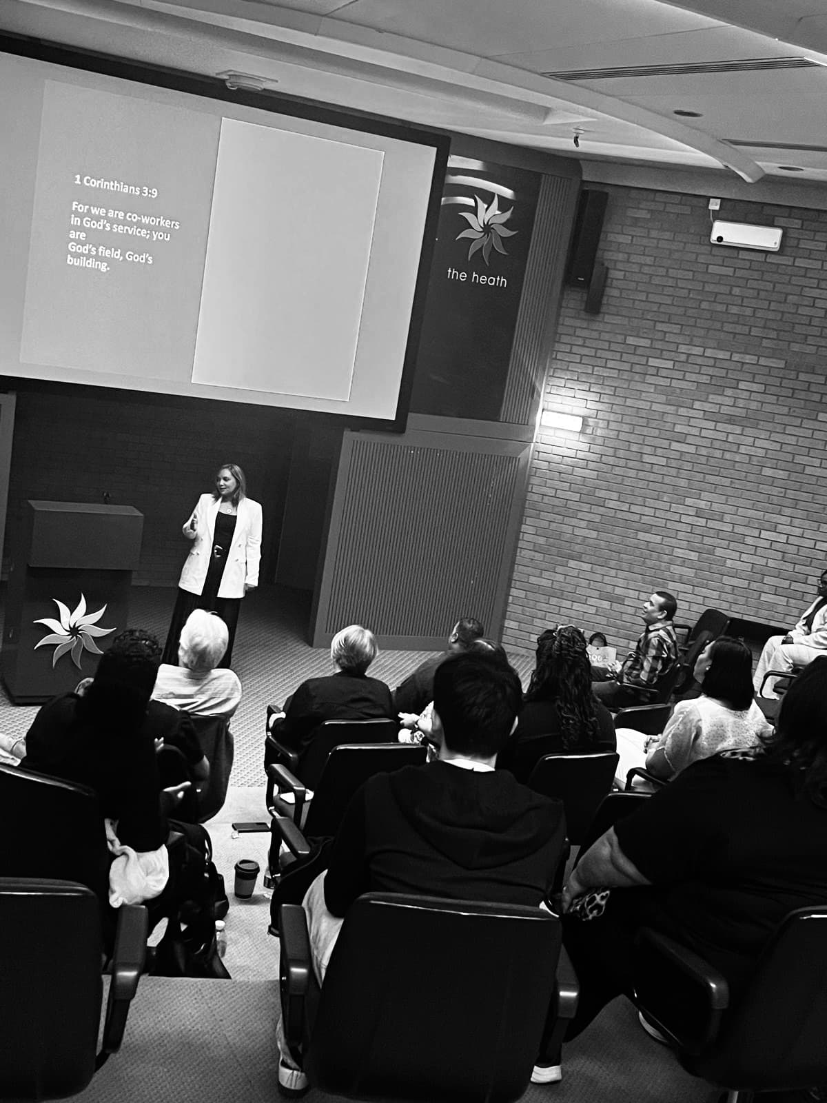 Woman in white blazer addresses an audience in a lecture hall with a projected verse.