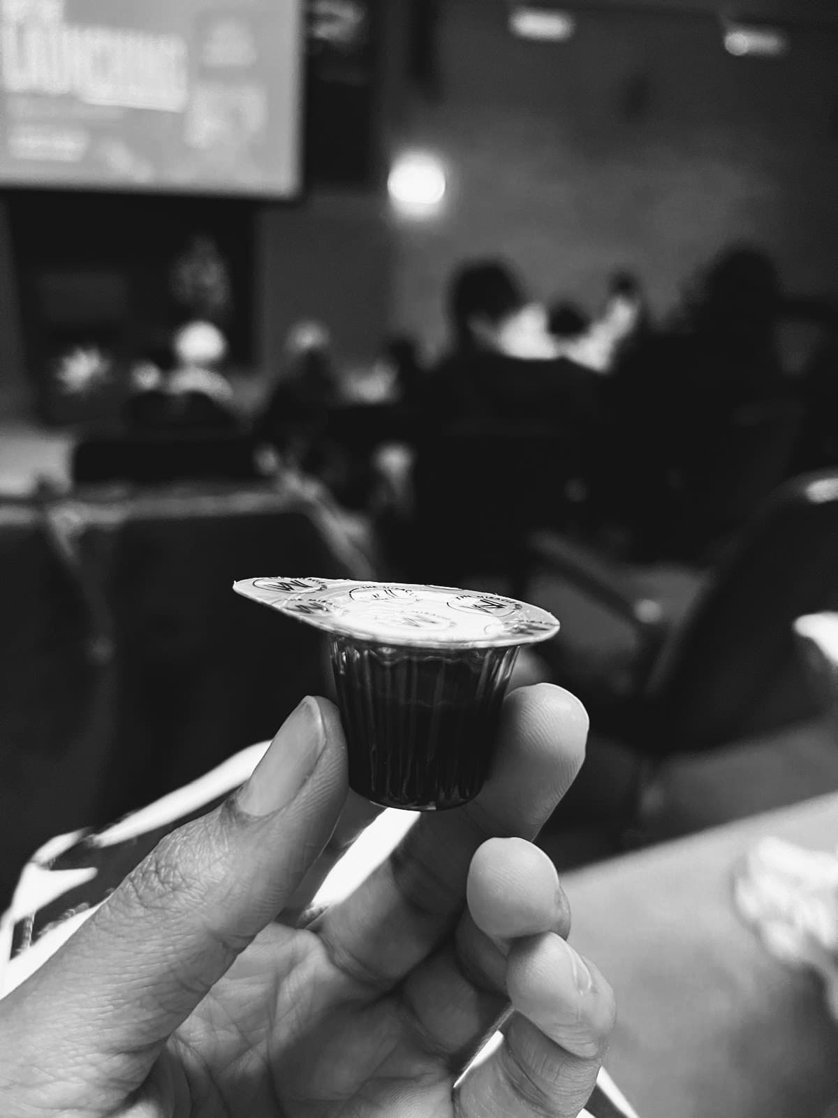 Hand holding a small, sealed communion cup in a blurred, black and white church auditorium.